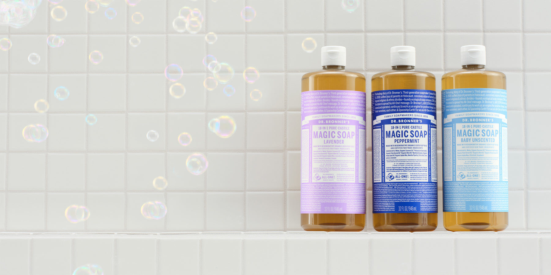 Three bottles of Dr. Bronner's Magic Soap on white tile with soap bubbles